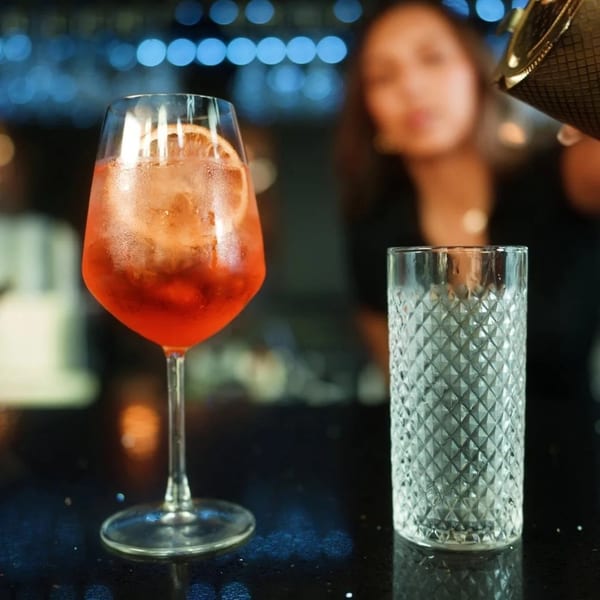 A bartender pours a drink into a tall glass next to a finished orange cocktail.