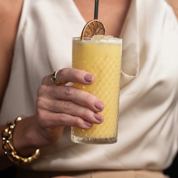 A person in a white top and gold jewelry holds a tall glass of yellow cocktail with a dried orange slice.