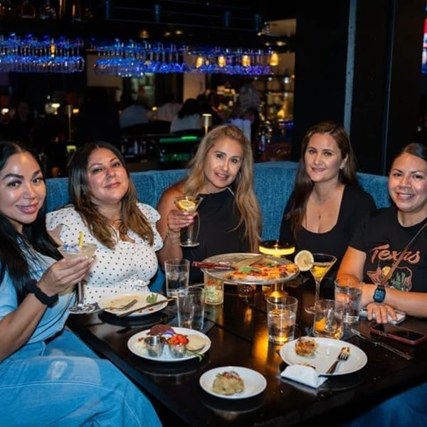 Five women enjoying drinks and food at a table in a dimly lit restaurant with blue booth seating.