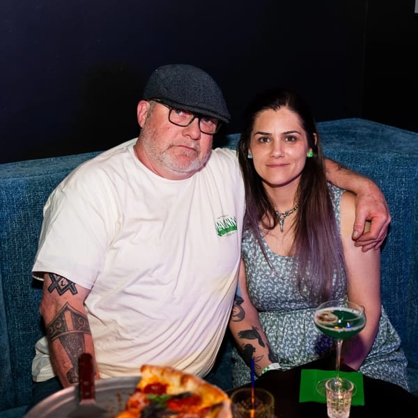 Man and woman sitting at a table with pizza and drinks, man's arm around woman.