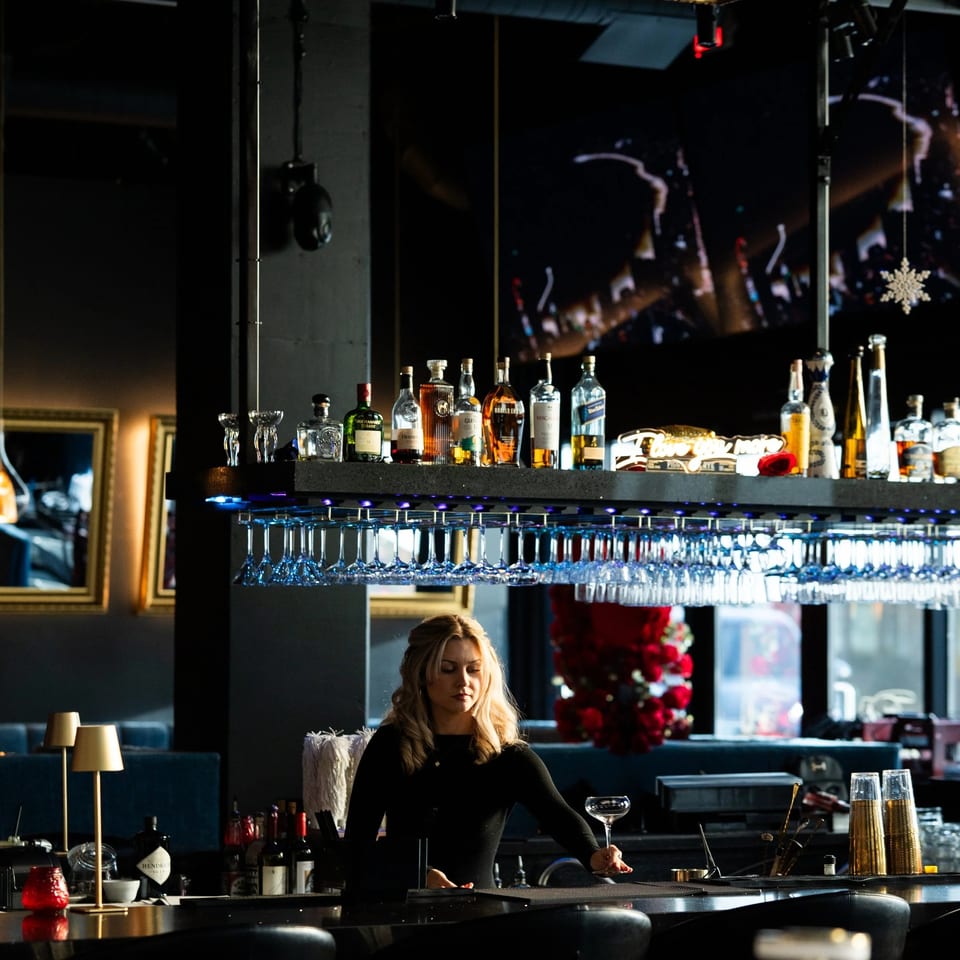 Blonde bartender in a black shirt holding a coupe glass behind a well-stocked bar.