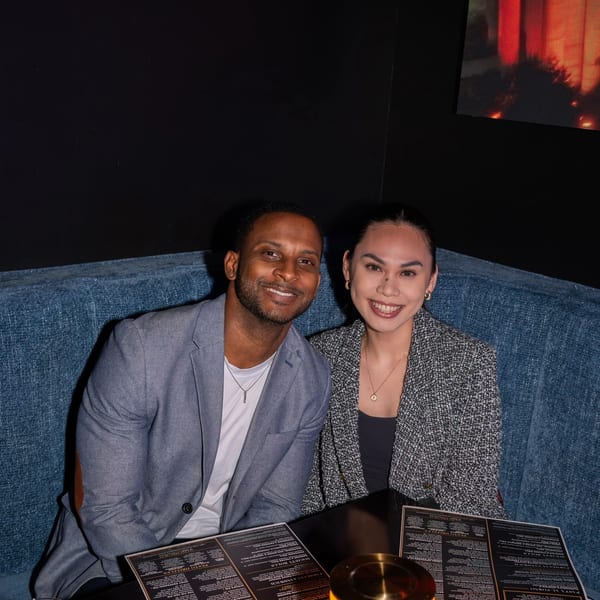 A smiling couple sits in a blue booth at a restaurant, with menus and drinks on the table.