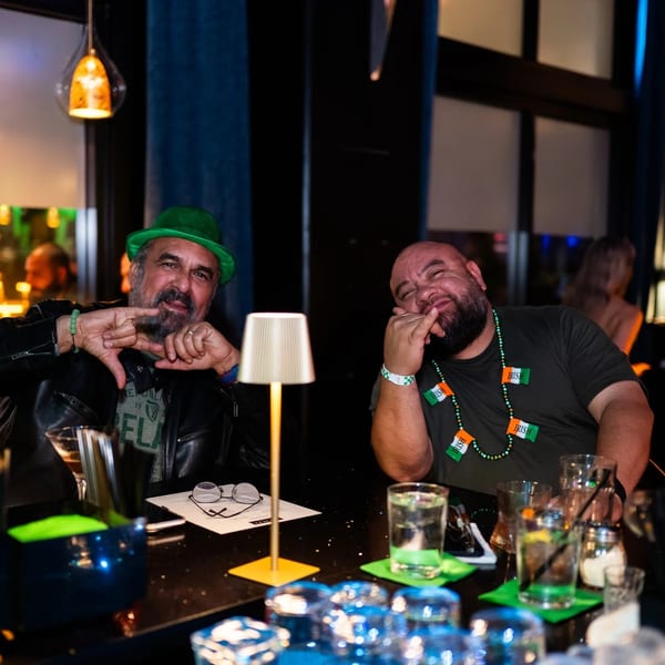 Two men in a dimly lit bar, one wearing a green hat, both smiling and gesturing.