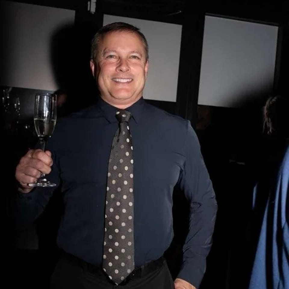 A smiling man in a dark shirt and polka-dot tie holds a glass of champagne.