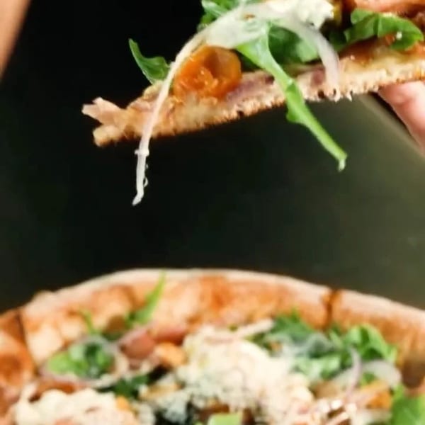 A hand holds up a slice of pizza topped with arugula, onions, and cheese, above a full pizza.