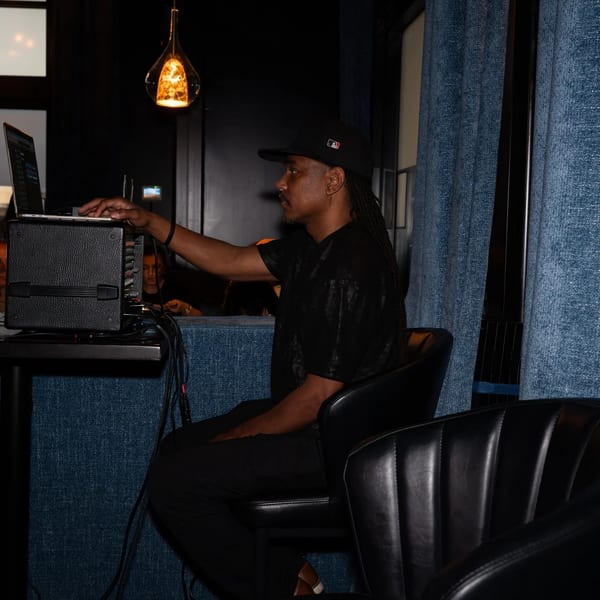 DJ in a black cap and shirt, operating a laptop and sound equipment in a dimly lit restaurant.