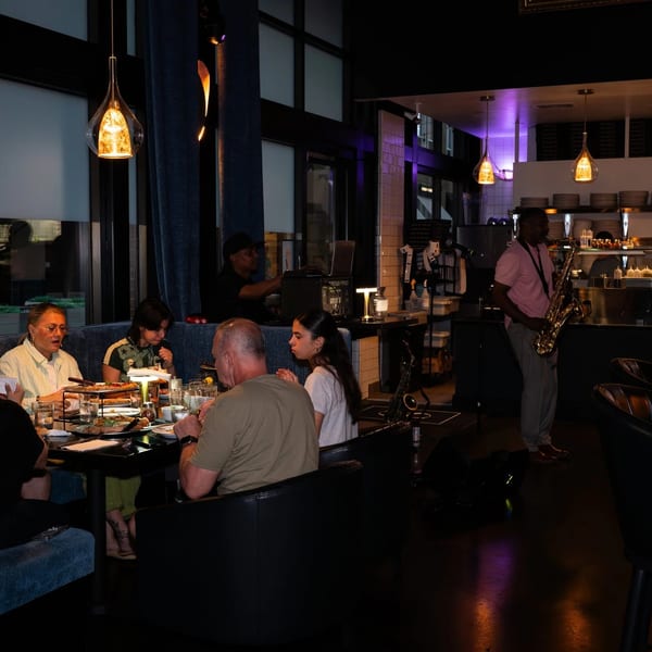 Diners at tables in a dimly lit restaurant with a saxophonist playing near the kitchen.