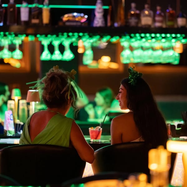 Two women with festive headwear enjoy drinks at a bar with green lighting and hanging glasses.