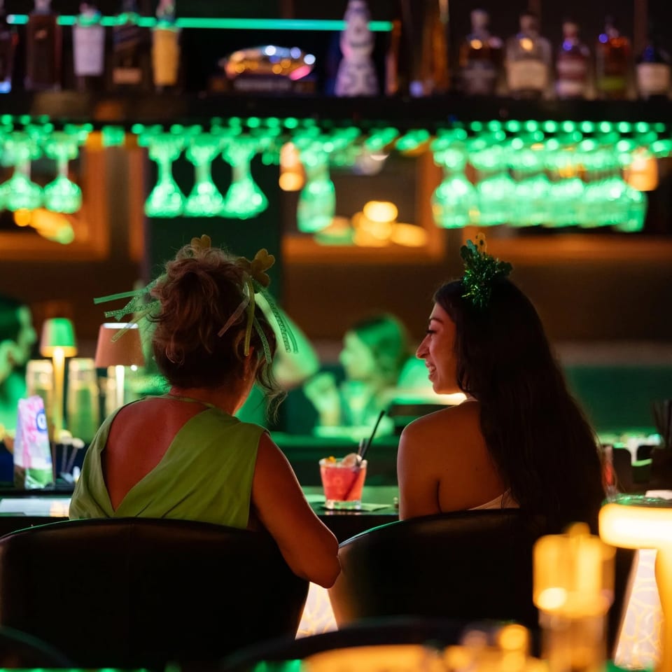 Two women with festive headwear enjoy drinks at a bar with green lighting and hanging glasses.