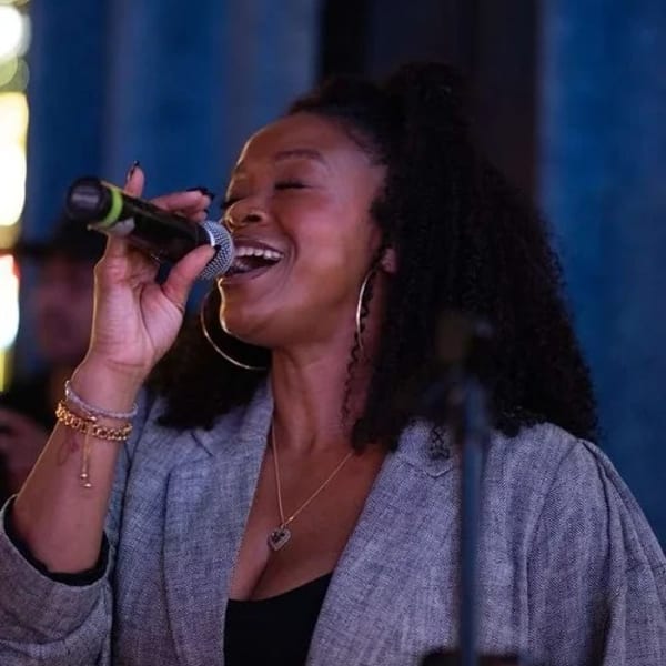 A woman with curly hair sings into a microphone, smiling with her eyes closed.