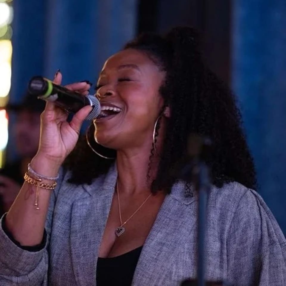 A woman with curly hair sings into a microphone, smiling with her eyes closed.