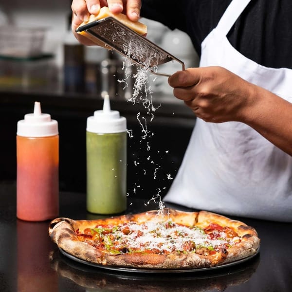 Chef grating fresh Parmesan cheese over a hot, delicious pizza with two sauce bottles nearby.