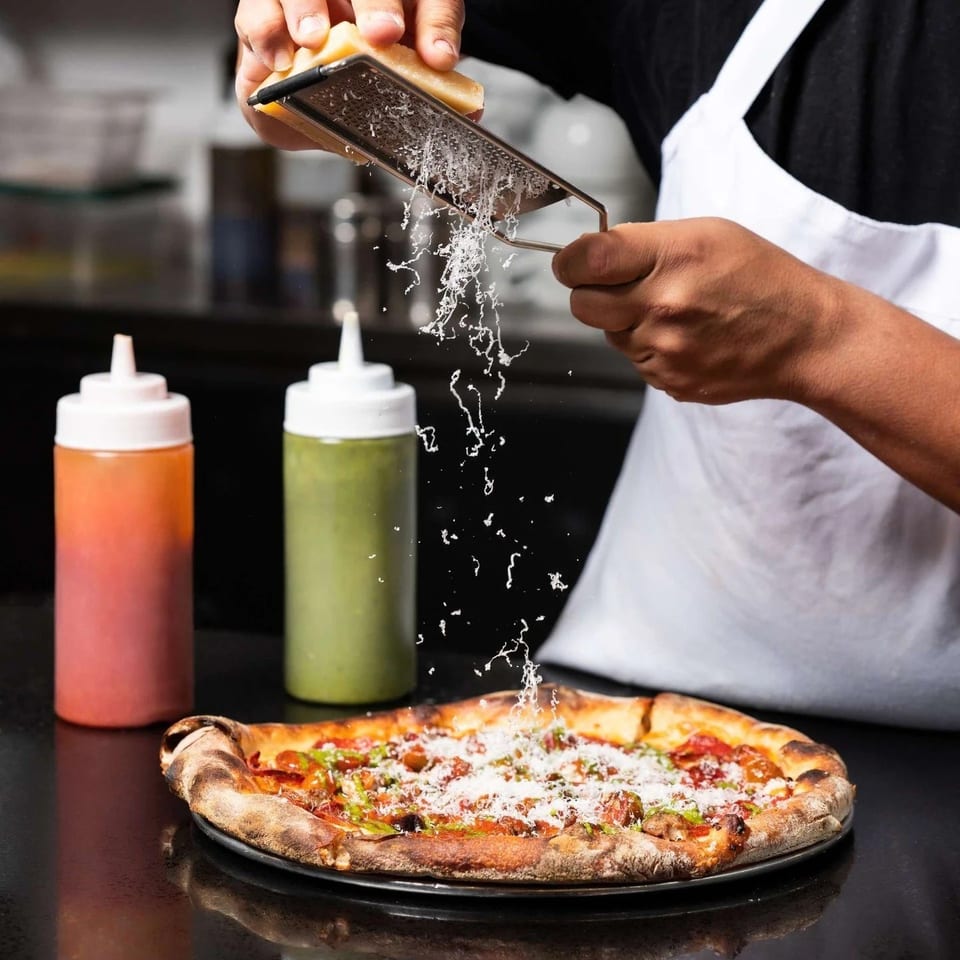 Chef grating fresh Parmesan cheese over a hot, delicious pizza with two sauce bottles nearby.