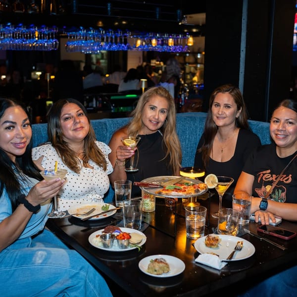 Five women smiling, enjoying drinks and food at a restaurant table with blue booth seating.