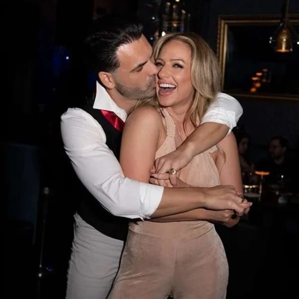Man in white shirt and red tie kissing a laughing woman in a beige jumpsuit on the cheek.