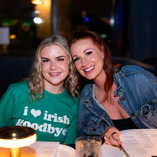 Two smiling women at a restaurant table, one in a green "I love Irish goodbye" shirt.