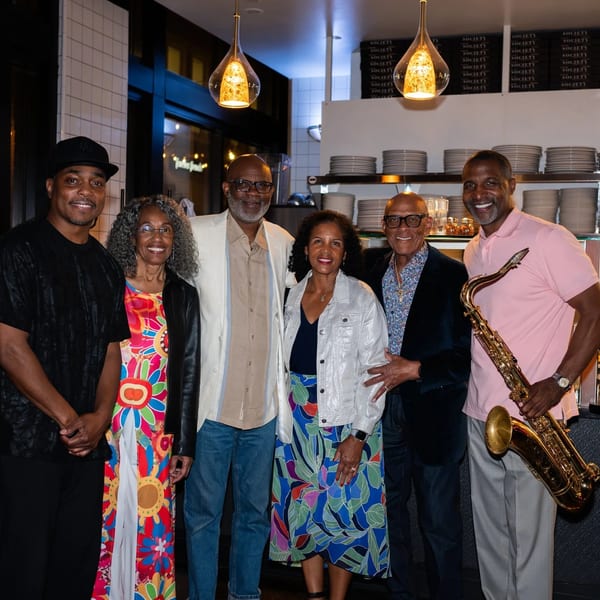 Six smiling people, including a man holding a saxophone, pose in a restaurant dining area.