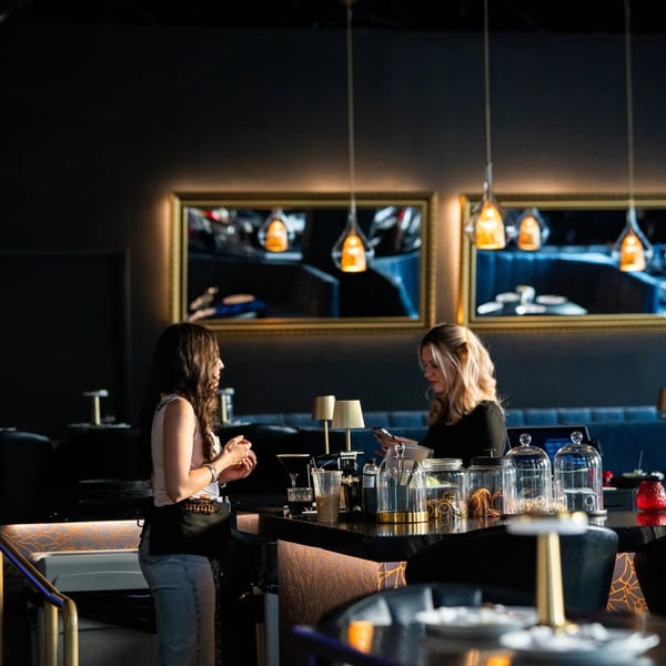 Two women interact at a dark, stylish bar with gold accents and hanging lights.