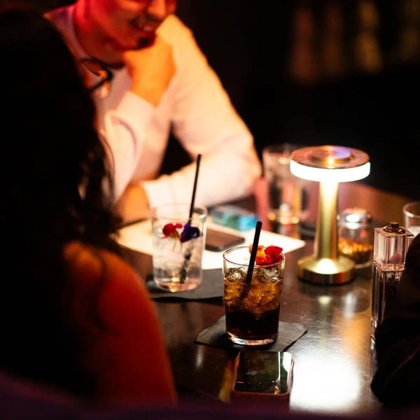 Two people at a dimly lit table with cocktails and a small lamp.