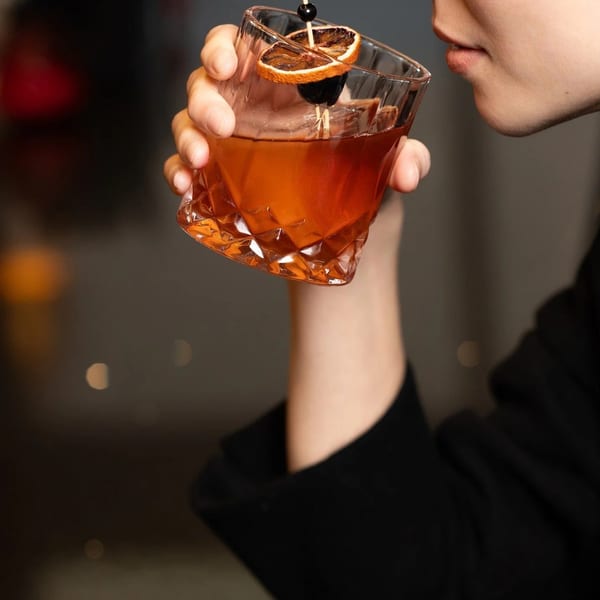 A person sips a cocktail garnished with dried orange and a cherry from a faceted glass.