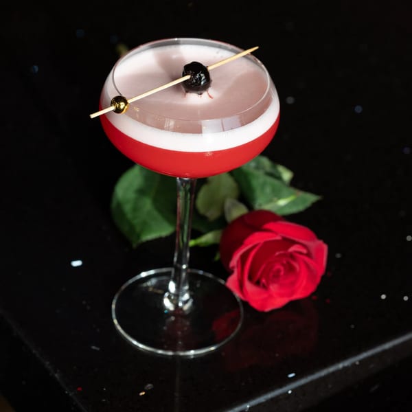 Red cocktail with white foam, garnished with a cherry and gold bead, next to a red rose.