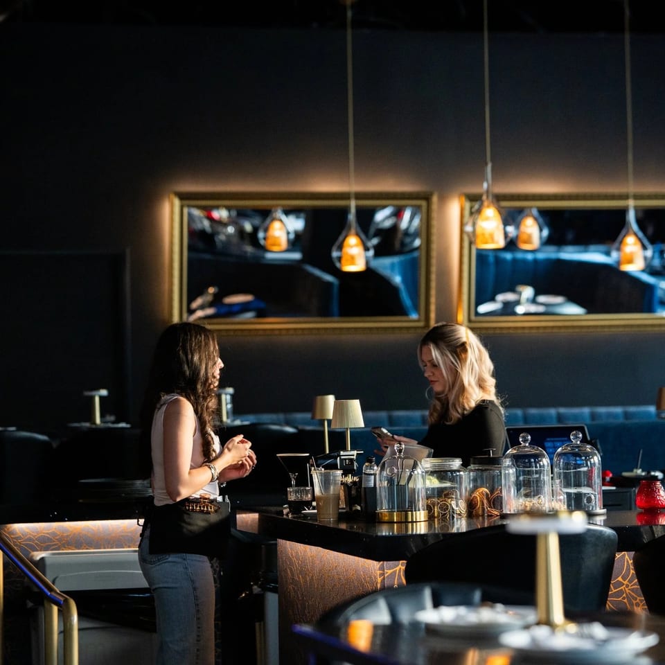Two women conversing at a dark, modern bar with gold accents and pendant lights.