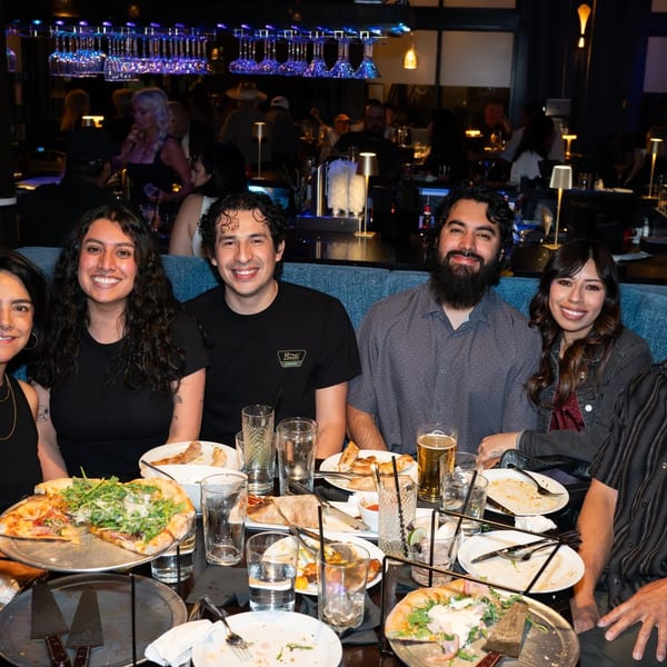 Six smiling people seated at a table with pizzas, drinks, and plates in a dimly lit restaurant.