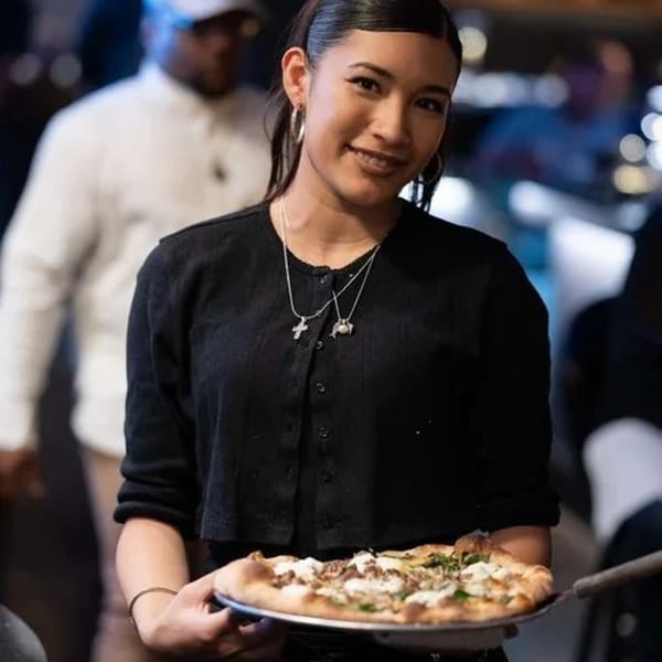 Smiling woman in black holding a freshly baked pizza on a metal peel, ready to serve.