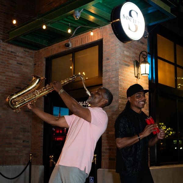 Two men performing music outside "Society" restaurant at night, one playing saxophone, the other holding shakers.