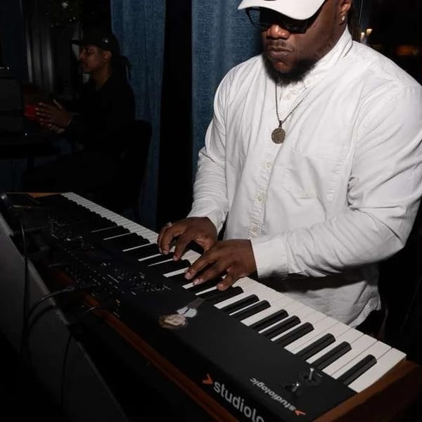 Musician in a white shirt and cap playing a Studiologic keyboard in a dimly lit venue.