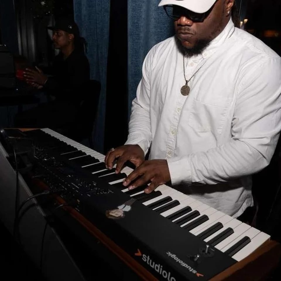 Musician in a white shirt and cap playing a Studiologic keyboard in a dimly lit venue.