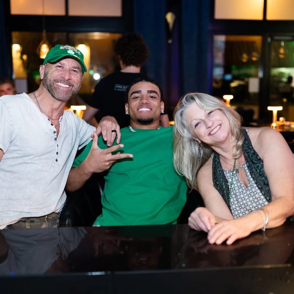 Three smiling people, two men and one woman, pose together at a dark bar or restaurant.