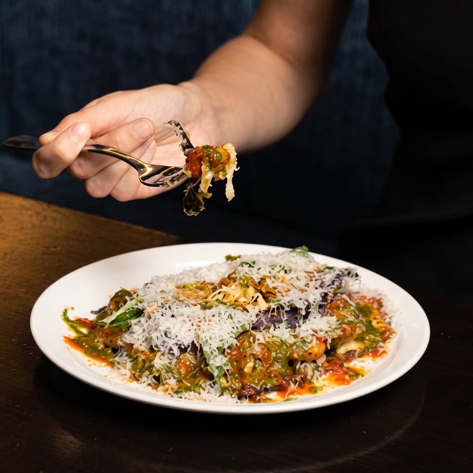 A hand holding a fork with a bite of pasta, covered in sauce and cheese, above a white plate.