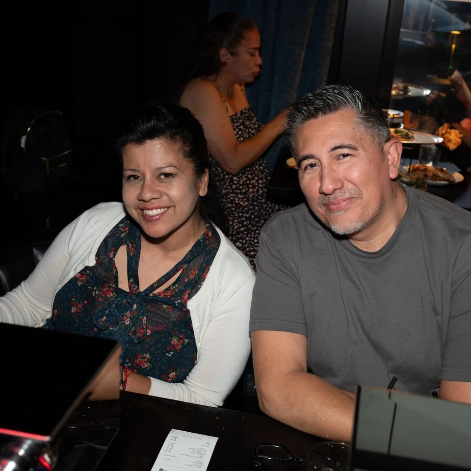 Two smiling people, a woman and a man, seated at a dark bar or table in a restaurant.