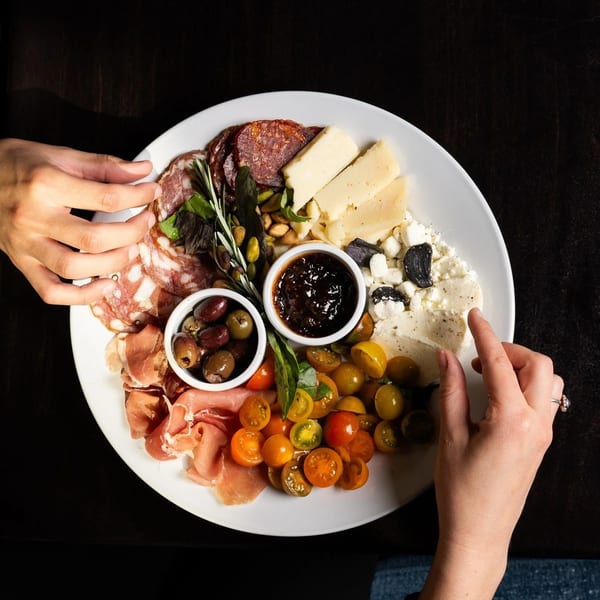 Two people sharing a charcuterie board with meats, cheeses, olives, tomatoes, and jam.